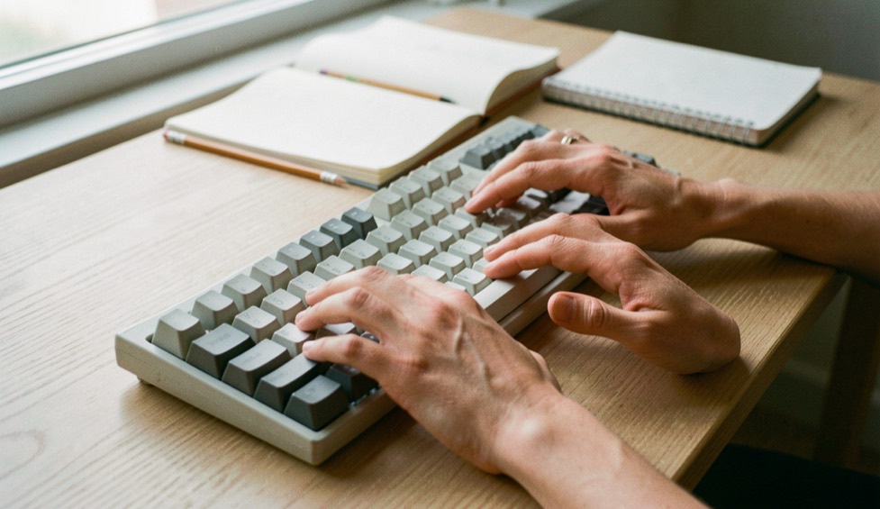 Two relaxed hands resting on the middle row of a mechanical keyboard with blank unlabelled keycaps, on a light oak desk beside a paper notebook and a pencil, soft morning light from a side window